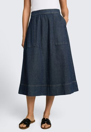 Donna che indossa una gonna midi in denim blu scuro con grandi tasche frontali, abbinata a sandali neri slip-on con punta aperta e una maglietta bianca.
