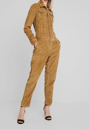 Kobieta modelka ubrana w beżowy sztruksowy kombinezon z długim rękawem, z przednimi kieszeniami, oraz czarne buty na obcasie z odkrytymi palcami, stojąca na jednolitym tle.