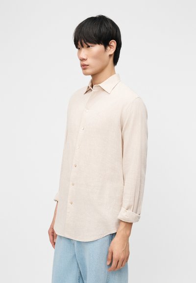 Νεαρός με ίσια μαύρα μαλλιά, φοράει μπεζ πουκάμισο με μακριά μανίκια και ανοιχτό μπλε τζιν, στέκεται μπροστά σε απλό φόντο.