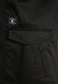 Musta kangas, jossa on vinokudonta, etutasku ja ommeltu merkkikilpi. Siistit linjat ja tyylikäs ulkonäkö.