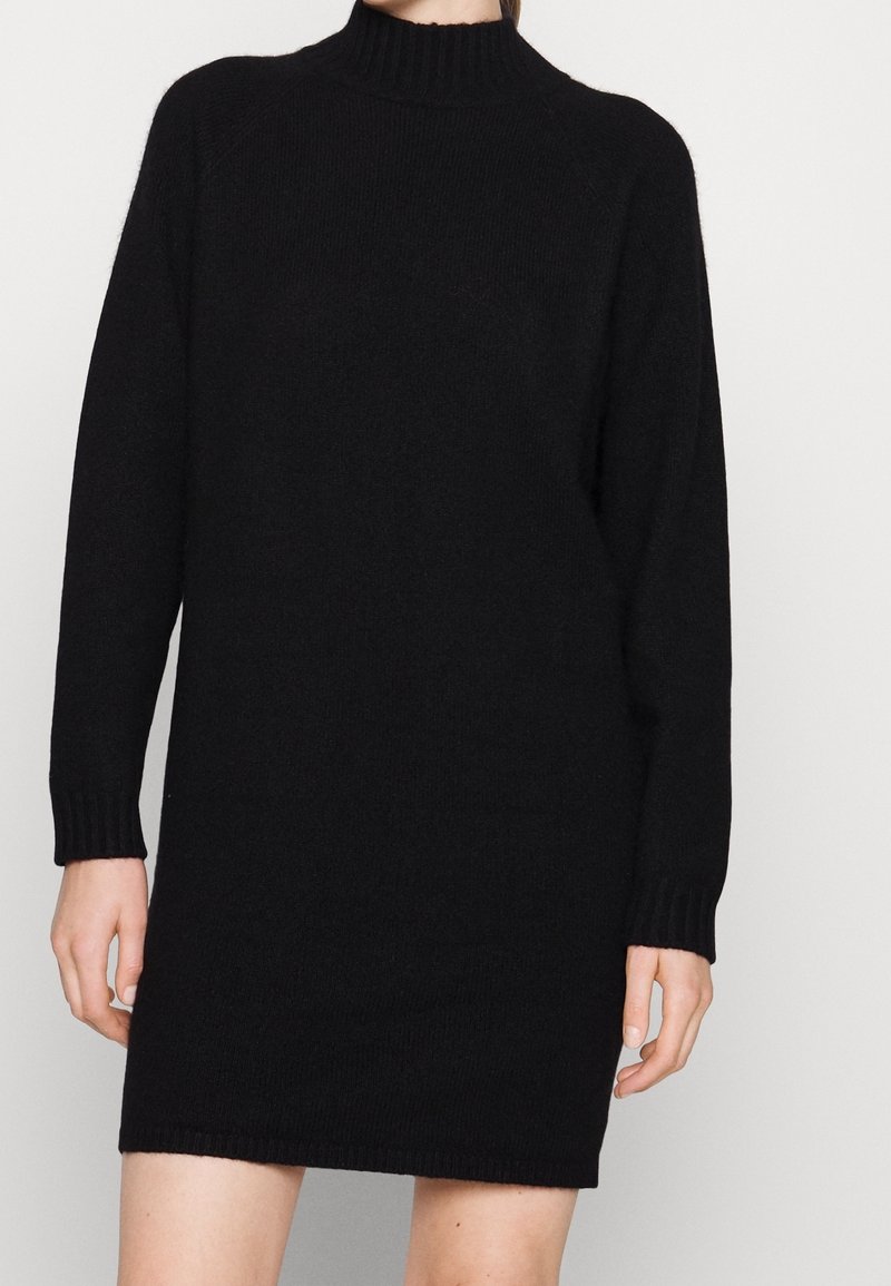 Femme portant une robe pull noire en maille à manches longues, avec un col rond montant et des poignets côtelés.