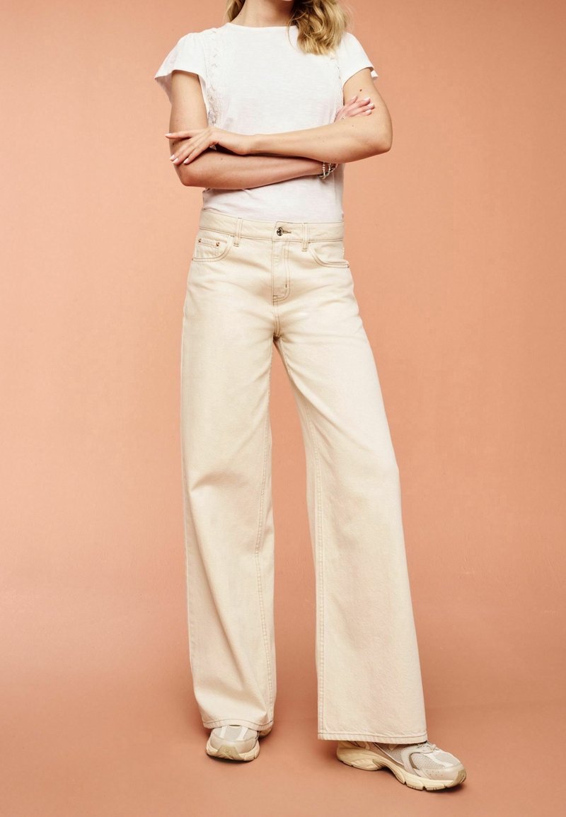 Femme portant un haut blanc à manches courtes, pantalon crème taille haute à jambes larges et baskets blanches, debout les bras croisés devant un fond pêche.