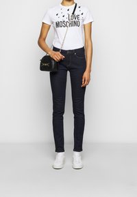 Žena v bílé triku "Love Moschino", tmavých skinny džínách, bílých teniskách, držící malou černou prošívanou kabelku s řetízkem.