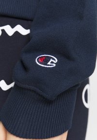 Poignet de sweat-shirt marine en tissu côtelé, avec un logo brodé blanc sur un patch rond avec des accents rouges et bleus.