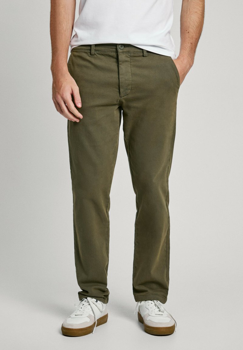 Pantalones de algodón verde oliva con un corte relajado, cierre de botón y bolsillos laterales, combinados con zapatillas blancas con suelas de color marrón claro.