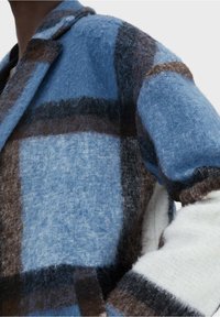 Abrigo de lana en patrón de cuadros azul y marrón, con un collar estructurado, textura suave y mangas en color crema contrastante.