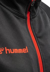 Veste noire texturée avec une fermeture éclair orange, comportant le logo "hummel" en orange sur le côté gauche.