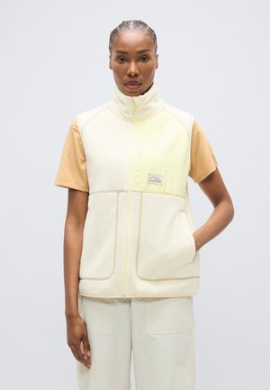 Fleecevest in crème en geel met een hoge kraag, twee voorzakken en contrasterende naden. Draagt over een bruin T-shirt met korte mouwen.