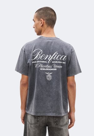 Mies seisoo selin, päällään haalistunut musta Benfica T-paita, jossa on valkoinen teksti ja tunnus, yhdistetty tummaan harmaaseen farkkuun.