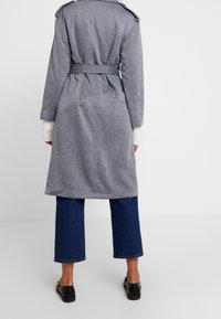 Šedý trenčkot s páskem, se strukturovaným límcem, texturovanou látkou a dlouhými rukávy, doplněný o zkrácené modré džíny a černé boty.