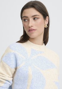 Vaaleanbeige neulepaita, jossa suuri sininen kukkakuvio, pyöreä kaula-aukko ja pehmeä pinta. Mallilla on kullanväriset renkaankorvakorut.