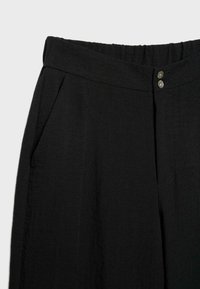 Mustat korkeavyötäröiset housut, joissa on joustava vyötärönauha, kaksi hopeanväristä nappia ja sivutaskut, esitettynä vyötäröstä reiden keskivaiheelle.