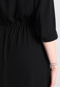 Černé šaty s elastickým pasem, nabíranými detaily a ohrnutými rukávy s knoflíkovými akcenty. Hladká textura látky, minimalistický design.