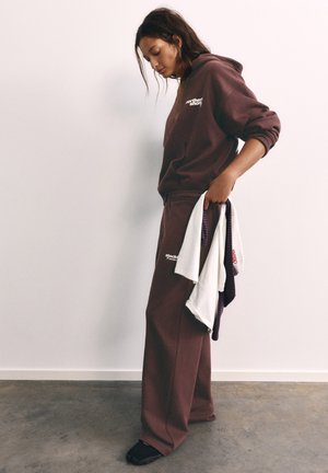 Femme portant un sweat à capuche marron et un pantalon large tenant des vêtements pliés, debout contre un mur blanc uni et un sol en béton.