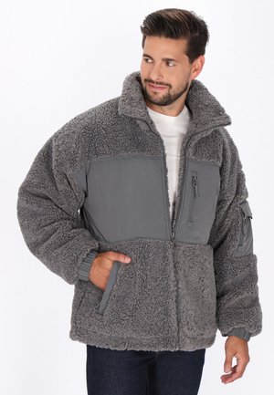 Mann in einer grauen, mit Reißverschluss versehenen Fleecejacke aus strukturiertem Material mit glatten Paneldetails, mit Händen in den Taschen, schaut zur Seite.