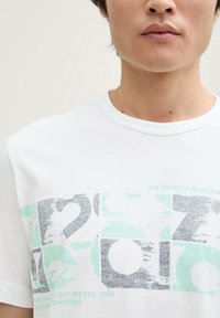 Hvid bomulds T-shirt med rund hals, prydet med abstrakt grå og mintgrøn tryk samt tekst i et moderne skrifttype hen over forsiden.