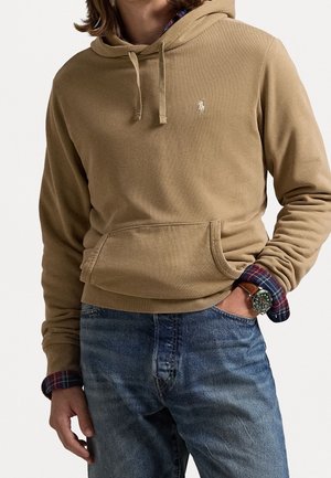 Beige hoodie i mjuk bomull med en känguruficka, ribbade muddar och en liten broderad logotyp på bröstet. Bärs över en rutmönstrad skjorta.