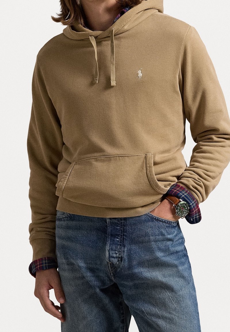 Felpa beige realizzata in morbido cotone, con tasca a marsupio, caratterizzata da polsini a coste e un piccolo logo ricamato sul petto. Indossata sopra una camicia a quadri.