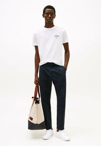 White cotton t-shirt with a small logo, navy pants, and white sneakers. Holding a beige and dark blue tote bag with brown handles.