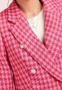 Kobieta w czerwono-różowej marynarce w wzór houndstooth z srebrnymi guzikami i nałożonymi złotymi naszyjnikami na białą bluzkę.