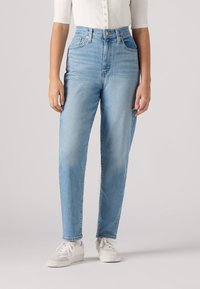 Personne portant un jean taille haute bleu clair, des baskets blanches et une chemise blanche côtelée à boutons, debout devant un fond uni.