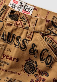 Pantalones cortos de tela color beige con texto impreso en negro y rojo y patrones florales. Presenta una etiqueta de Levi's y detalle de botón metálico.