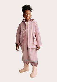 Růžová nepromokavá bunda s kapucí, doplněná o odpovídající kalhoty a krémové gumové boty. Má elastické manžety a reflexní prvky.