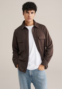 Barna hosszú ujjú ing két elülső zsebbekkel, gombos záródással, puha textúrával, fehér póló fölött viselve, világoskék farmernadrággal párosítva.