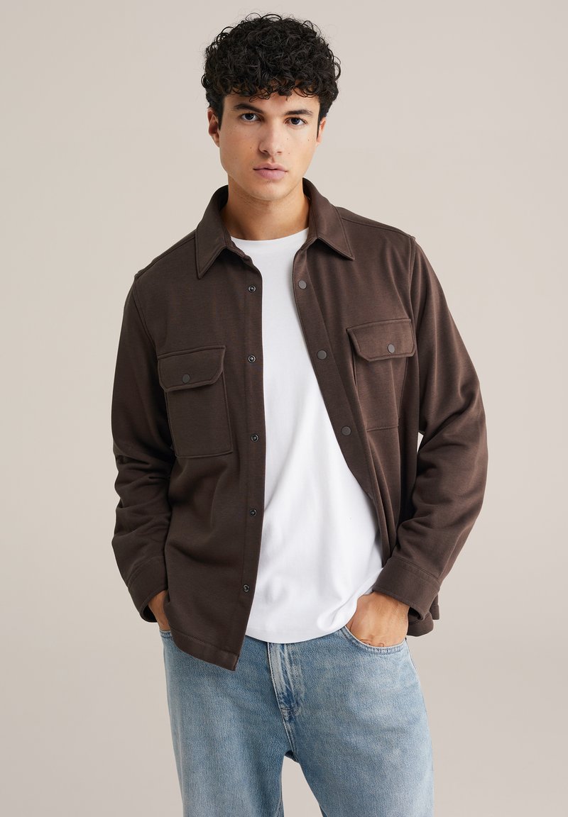 Barna hosszú ujjú ing két elülső zsebbekkel, gombos záródással, puha textúrával, fehér póló fölött viselve, világoskék farmernadrággal párosítva.