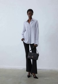 Camisa de manga longa riscada em branco e azul-marinho, combinada com calças pretas amplas e sapatos de salto agudo. O modelo segura uma bolsa preta texturizada.