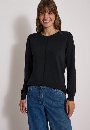 Femme souriante portant un pull noir à manches longues avec une couture au centre et un jean bleu sur un fond uni.