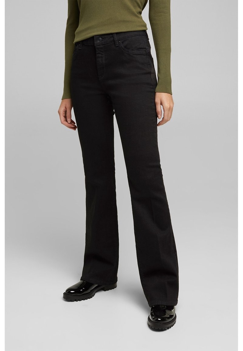Personne portant un jean noir évasé, un haut olive à manches longues et des chaussures noires brillantes à lacets, debout devant un fond gris clair uni.