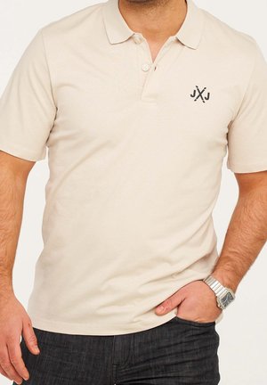 Mann trägt ein beiges, kurzärmeliges Polohemd mit einem kleinen schwarzen Logo auf der Brust und eine silberne Armbanduhr, die Hand in der Tasche.