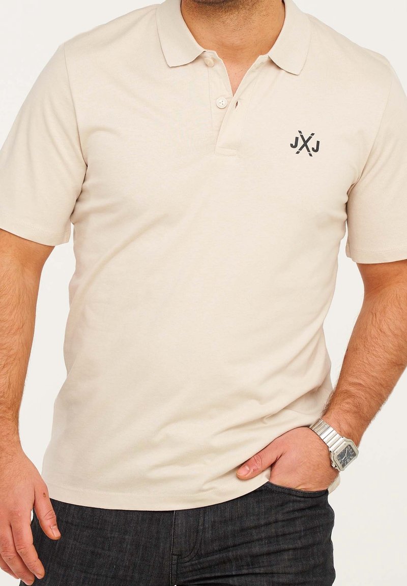 Homme portant un polo beige à manches courtes avec un petit logo noir sur la poitrine et une montre-bracelet argentée, la main dans la poche.