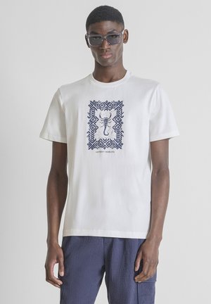 Joven vestido con gafas de sol y una camiseta blanca con un diseño azul de escorpión y pantalones texturizados azul marino, de pie frente a un fondo liso.