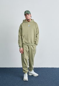 Oliven grøn hoodie- og sweatpants-sæt i blødt stof med et centralt logo-patch. Afsluttet med hvide sneakers og en grøn kasket.