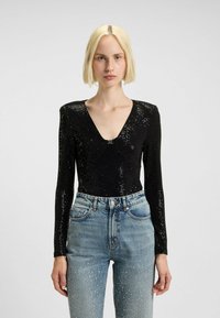 Blonde vrouw in een zwart met pailletten bedekt lang mouwtop met een diepe V-hals en lichtblauwe hoogwaistjeans met kristalversieringen.
