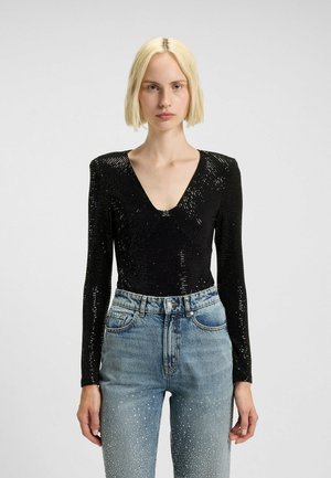 Donna bionda che indossa un top lungo a maniche nere con paillettes e scollatura a V profonda, abbinato a jeans a vita alta di colore azzurro chiaro con decorazioni di cristallo.