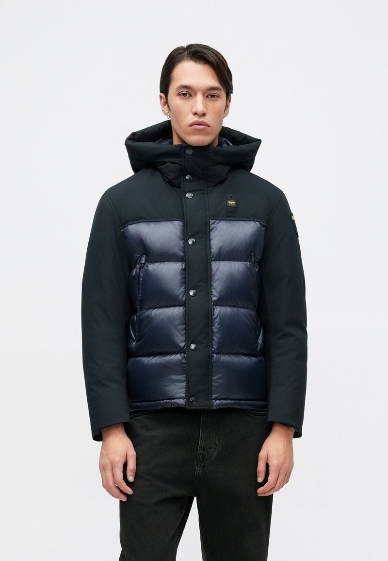 Doudoune noire avec un devant matelassé bleu marine foncé, capuche et fermetures à boutons. Finition lisse avec poches visibles et accent logo.