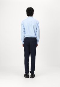 Hellblaues Langarmhemd mit geradem Schnitt, kombiniert mit dunkelblauen Hosen und schwarzen Schuhen, von hinten vor einem weißen Hintergrund gezeigt.