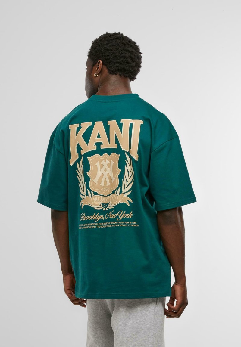 Tricou supradimensionat verde din bumbac, cu un design grafic auriu pe spate care include textul "KANI" și "Brooklyn, New York".