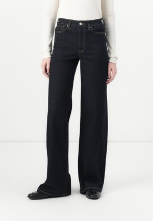 Osoba nosząca ciemne dżinsy z wysokim stanem i szerokimi nogawkami, połączone z czarnymi butami oraz białą bluzką z długim rękawem, stojąca na jednolitym białym tle.