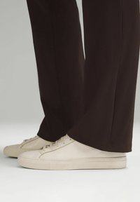 Baskets en cuir blanc portées avec un pantalon large marron foncé, debout sur une surface gris clair.