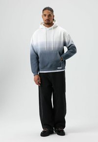 Heren hoodie met verloopkleur van wit naar donkerblauw, voorzien van een voorzak en een capuchon met trekkoord, gecombineerd met losse zwarte jeans en grove schoenen.