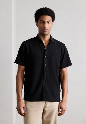 Moški s kratkimi kodrastimi lasmi, oblečen v črno kratkoročno srajco z gumbi in bež hlače, stoji pred enobarvnim svetlim ozadjem.