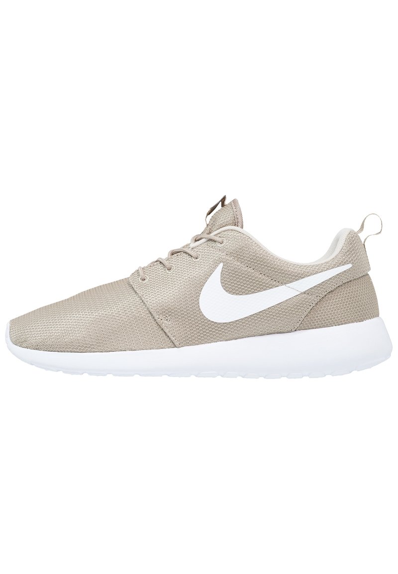 Ljusbeige Nike-sko med vit swoosh-logotyp, ovandel i mesh, snörning och tjock vit sula, visad från sidan mot en vit bakgrund.