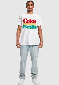 Bílý bavlněný tričko s červeným, žlutým a zeleným textem: "Coke always fresh." V kombinaci se světle modrými džíny a bílými teniskami.