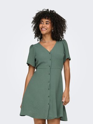 Donna con capelli ricci sorridente, che indossa un vestito verde con bottoni e maniche corte e una collana d'argento, in piedi davanti a uno sfondo chiaro e uniforme.