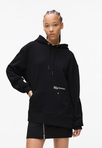 Black oversized hoodie made of soft fabric, featuring a front pocket and embroidered logo detail. Paired with a black skirt with a zipper.