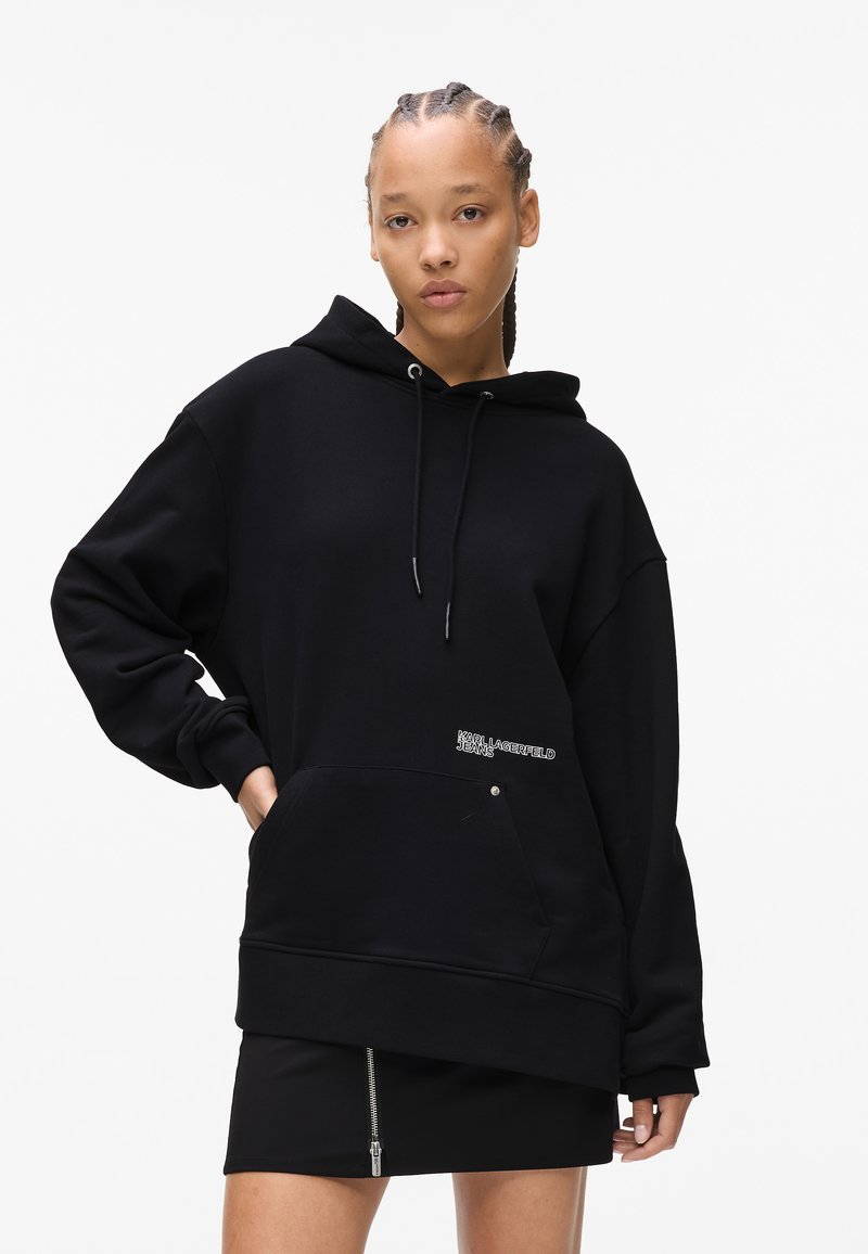 Black oversized hoodie made of soft fabric, featuring a front pocket and embroidered logo detail. Paired with a black skirt with a zipper.